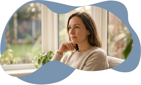 A thoughtful woman looking out a window, framed by a soft blue organic shape, illustrating the search
for clinical certainty and a foundational roadmap through a formal adult autism assessment.